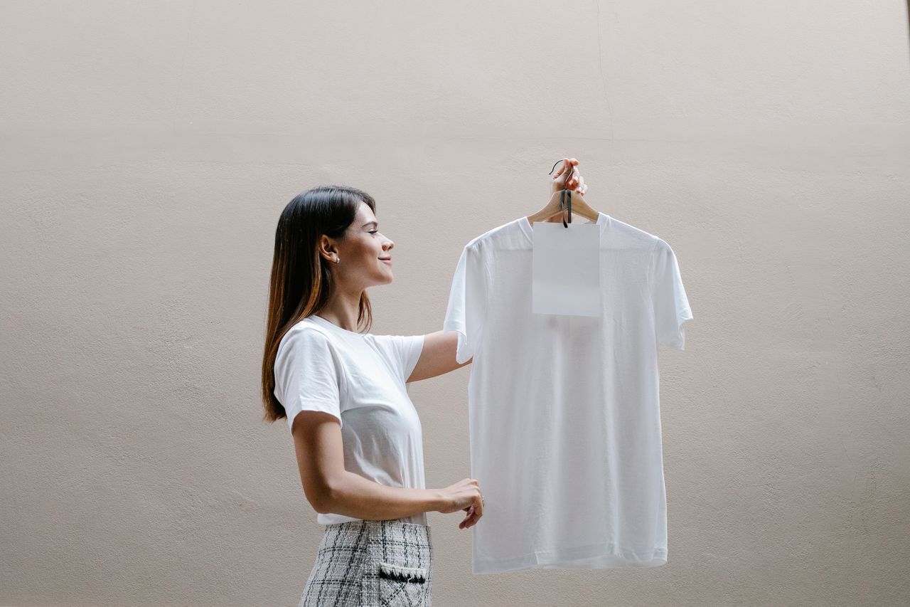 The Art of Drawing Readers In: Your attractive post title goes here Side view of positive young female designer in stylish clothes holding hanger with white t shirt and blank paper sheet and smiling against gray background